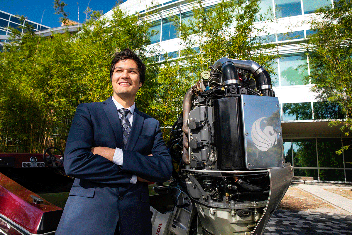 A student stands in a suit with his arms crossed in front of a giant engine on the back of a boat with the 첥 Pegasus logo.