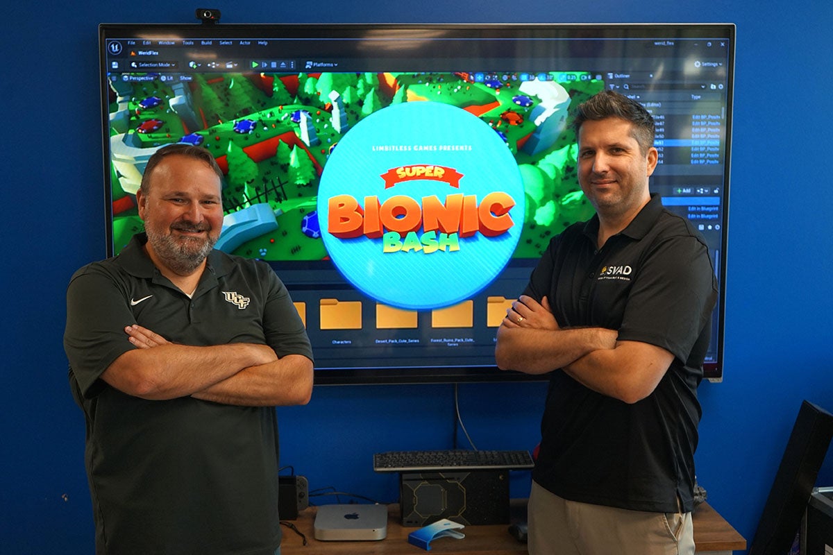 첥 Associate Professors Peter Smith and Matt Dombrowski stand in front of an screenshot of their new game Super Bionic Bash that is designed to help people learn to use their prosthetic limb.